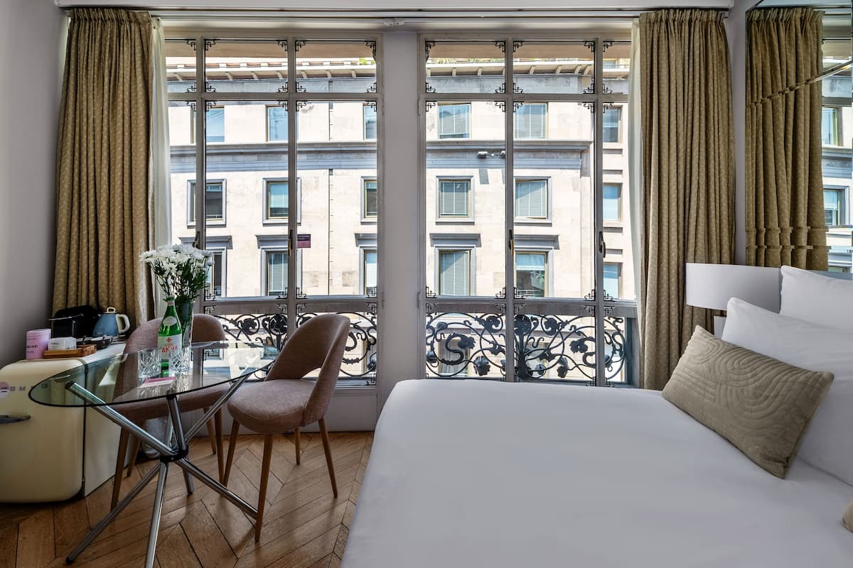Hotellihuone, jossa on suuri sänky, lasinen pöytä tuoleineen, näkymä rakennuksiin suurista ikkunoista sekä verhot.