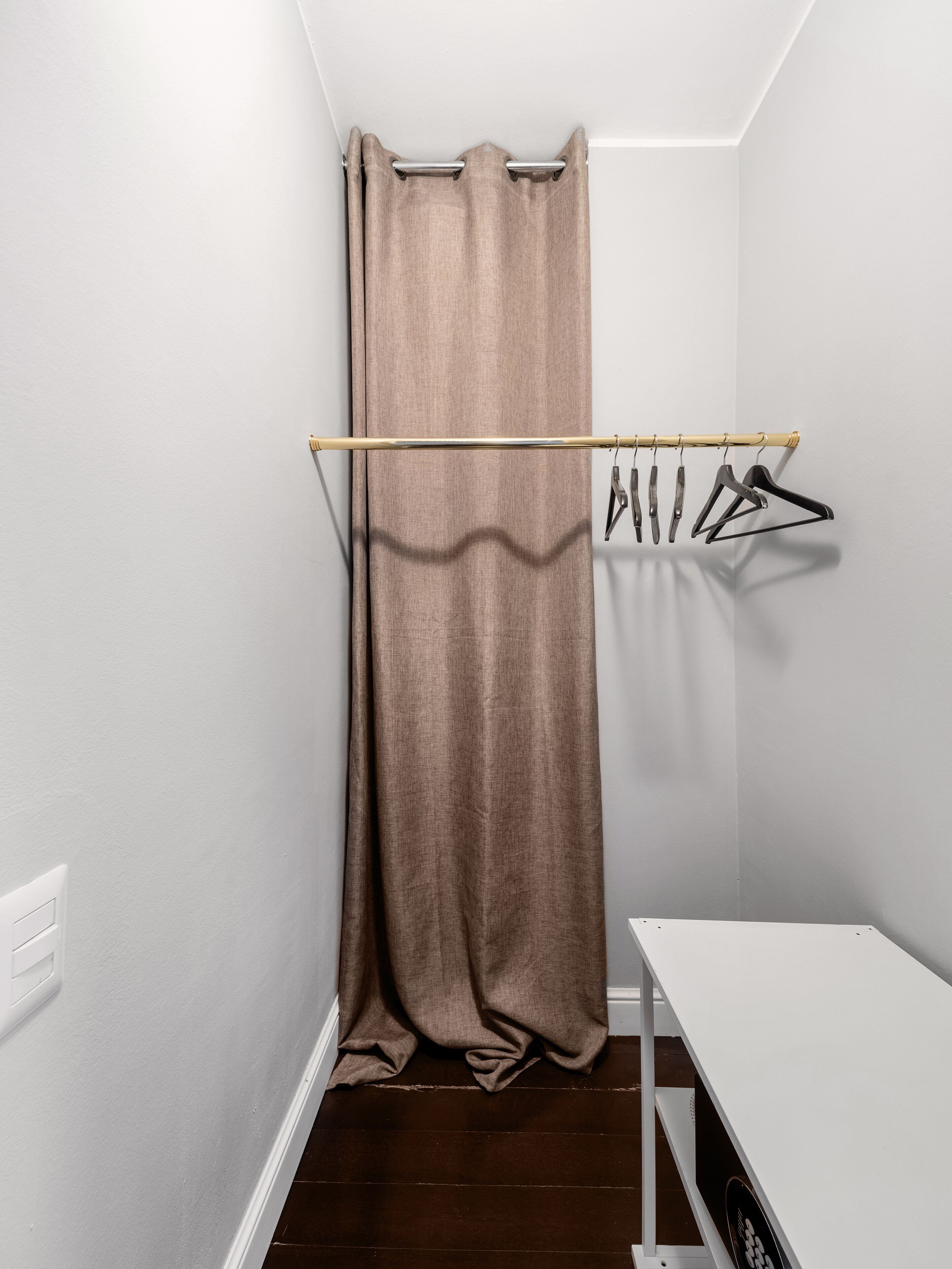 A small, narrow room with a wooden floor, a white wall, and a wooden clothes rack with hangers.