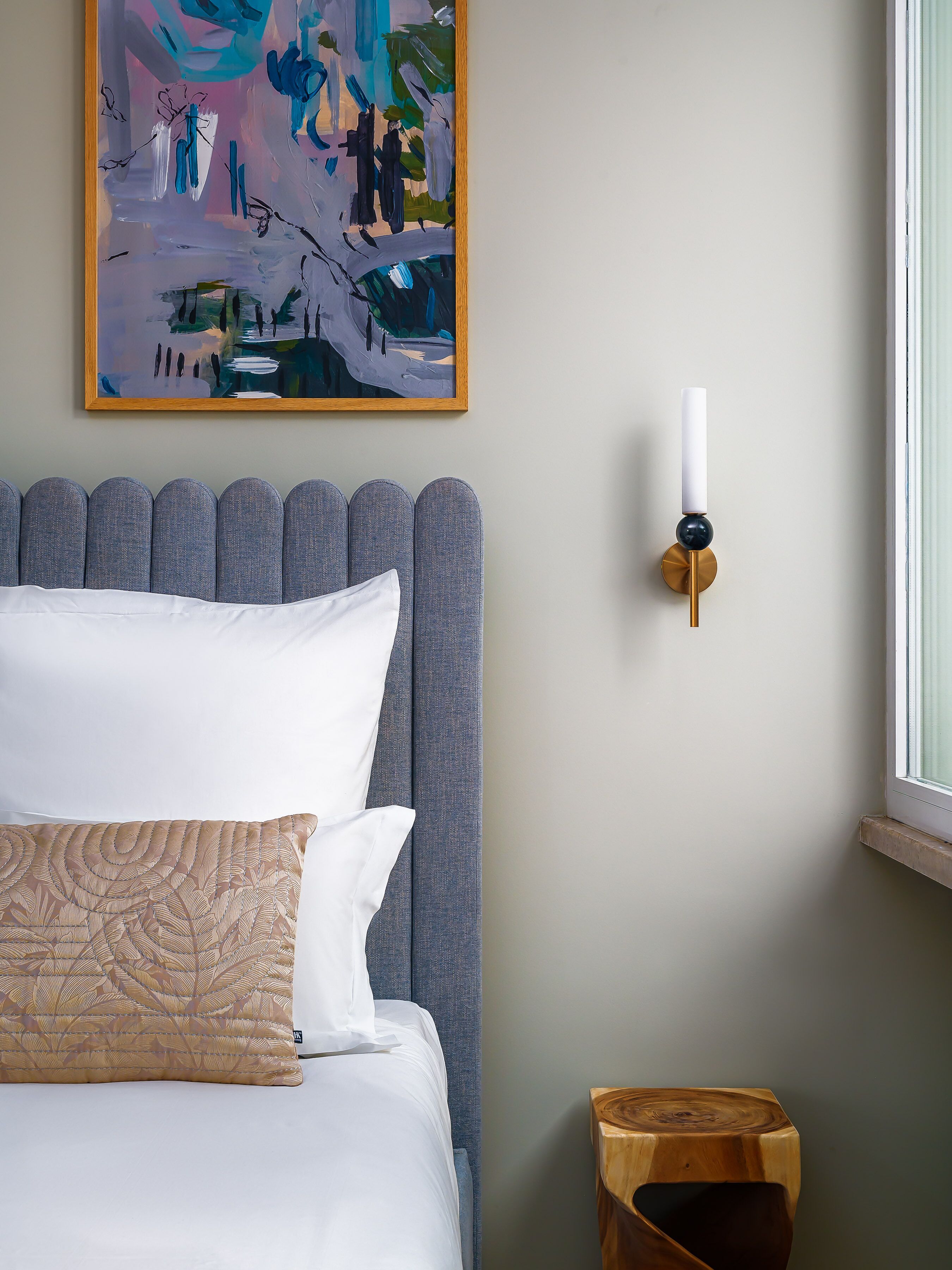 Un dormitorio con cabecera tapizada gris, colcha blanca y un cojín decorativo estampado. Encima de la cama hay un cuadro abstracto enmarcado, y a la derecha se ve una lámpara de pared.