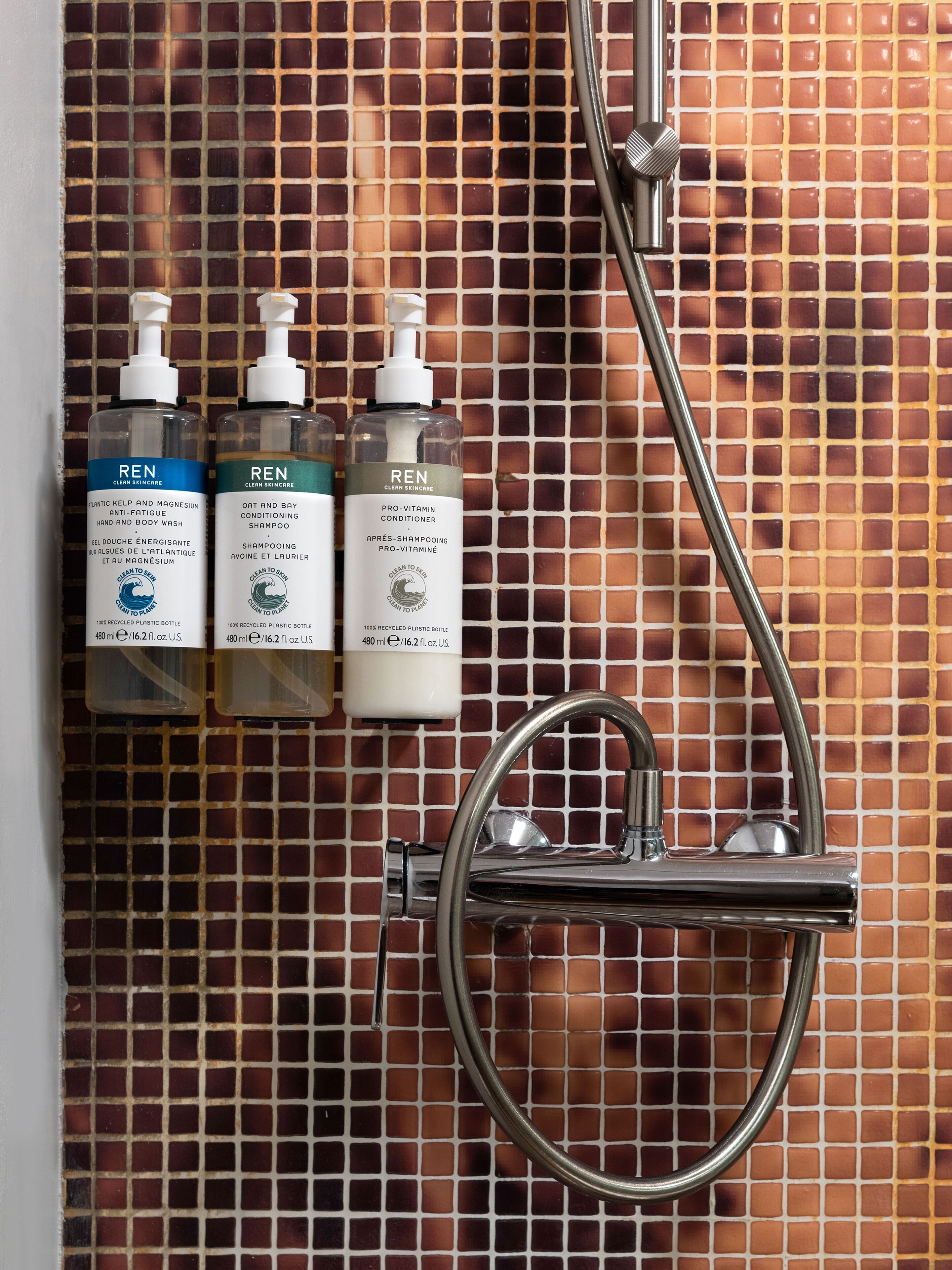 Un baño con pared de azulejos, tres botellas de productos REN para el cuidado de la piel y un cabezal de ducha cromado.