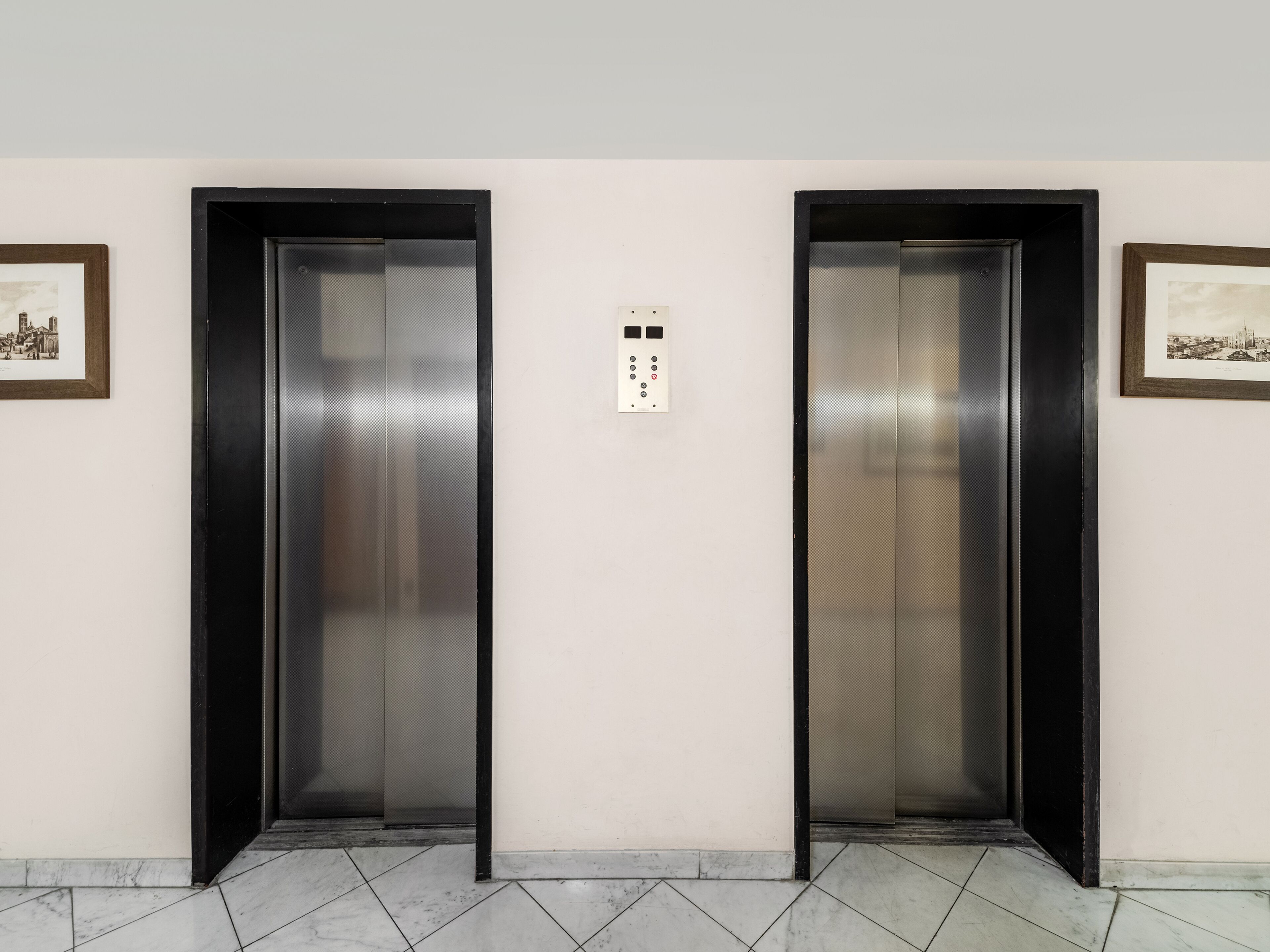 Two elevator doors in a hallway with a tiled floor and two framed pictures on the walls.