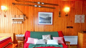 A cozy room with a wooden interior, a bed with red bedding, a small table with a basket, and a wall-mounted shelf with chocolate jars.