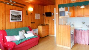 A cozy cabin interior with a wooden floor, a red and green sofa, a wooden dresser, a television mounted on the wall, and a kitchen area with a sink and cabinets.