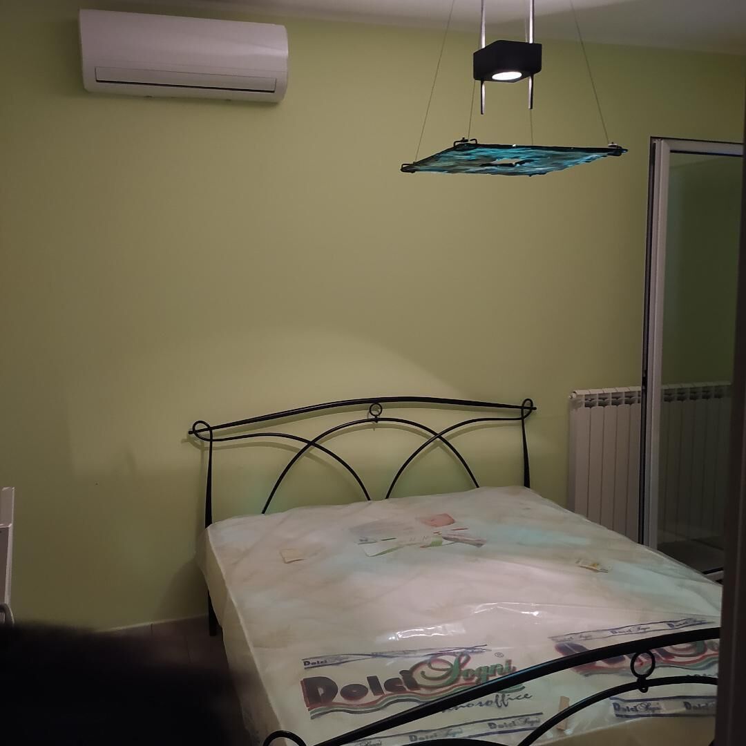 Un dormitorio con estructura de cama metálica, una cama individual con colchón blanco, aire acondicionado de pared y lámpara colgante.