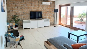 A modern living room with a stone wall, a flat-screen TV, a white entertainment unit, a floor lamp, a sofa, a coffee table, and a sliding glass door leading to an outdoor area. - House by the sea (Cefalù)