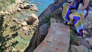 A woman sitting on a stone ledge overlooking a rocky coastline with clear blue water. - House by the sea (Cefalù)