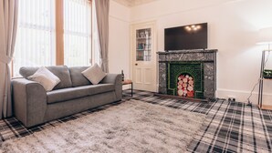 A living room with a grey sofa, a fireplace, a television, and a checkered rug. - Eden Cottage- Quiet City Base (Dundee)