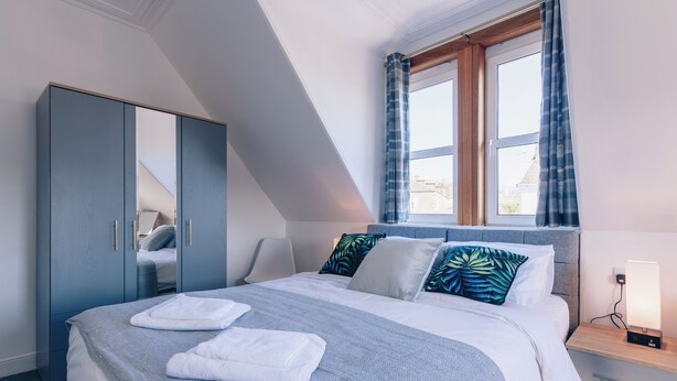 A modern bedroom with a bed, a wardrobe, a bedside table with a lamp, and a window with curtains. - Eden Cottage- Quiet City Base (Dundee)