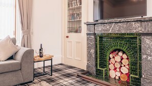 A living room with a fireplace, a sofa, a table with a bottle and glasses, and a bookshelf. - Eden Cottage- Quiet City Base (Dundee)
