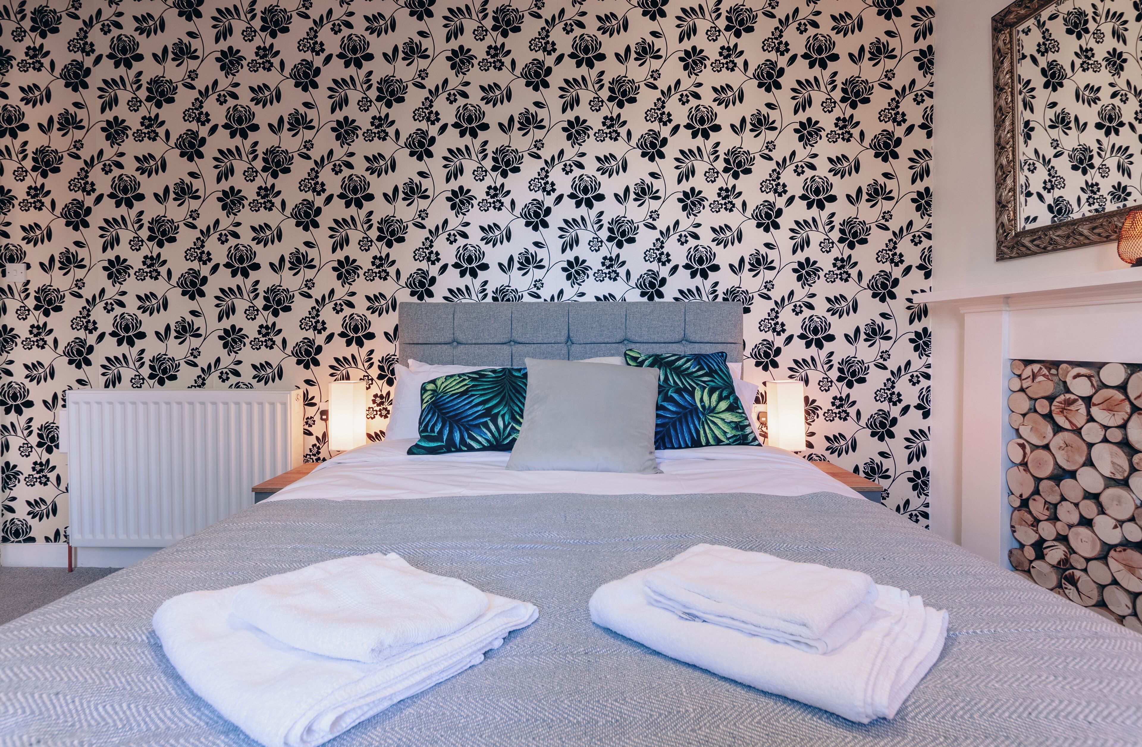 A neatly made bed with folded towels, a headboard, and floral wallpaper.