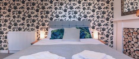 A neatly made bed with folded towels, a headboard, and floral wallpaper.