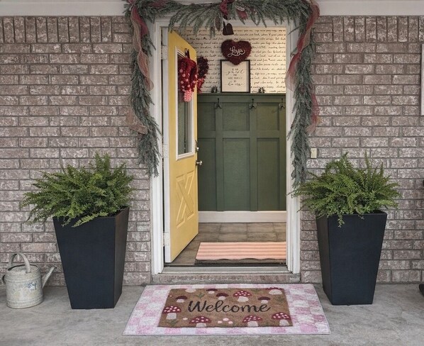 A brick exterior with a green door, two potted plants, a welcome mat, and a sign above the door.