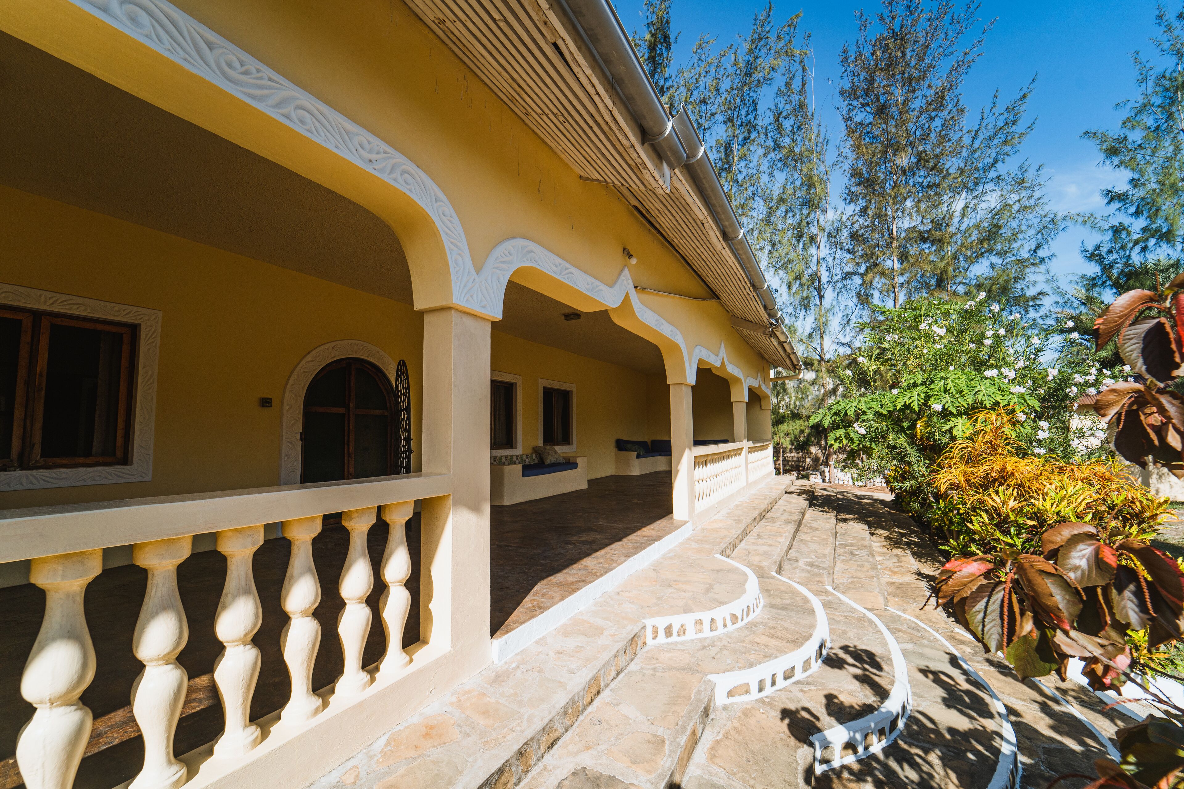 A building with a yellow facade, white decorative arches, and a stone pathway leading to a covered area with seating.