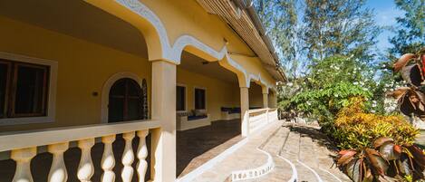 A building with a yellow facade, white decorative arches, and a stone pathway leading to a covered area with seating.