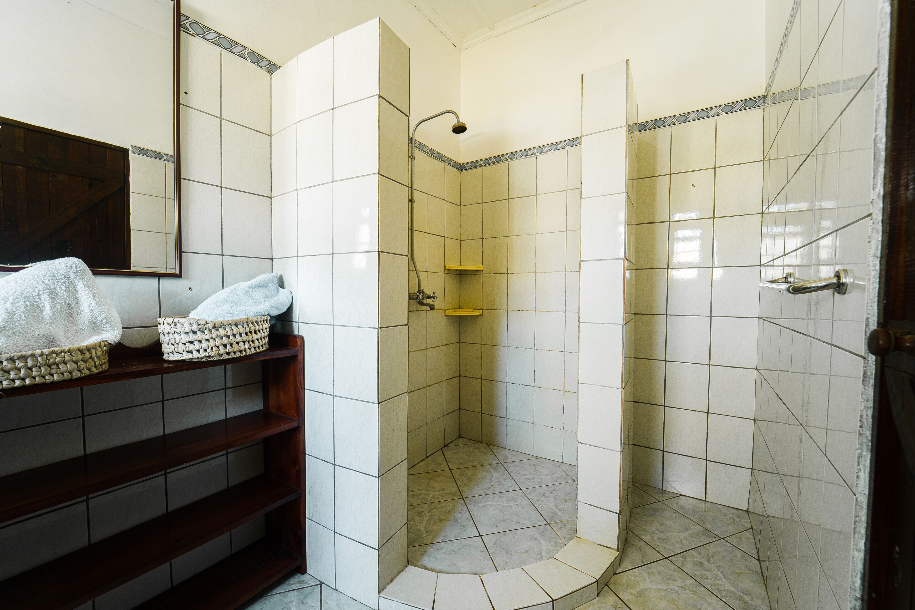 A bathroom with a shower, tiled walls, a wooden shelf with baskets, and a mirror.