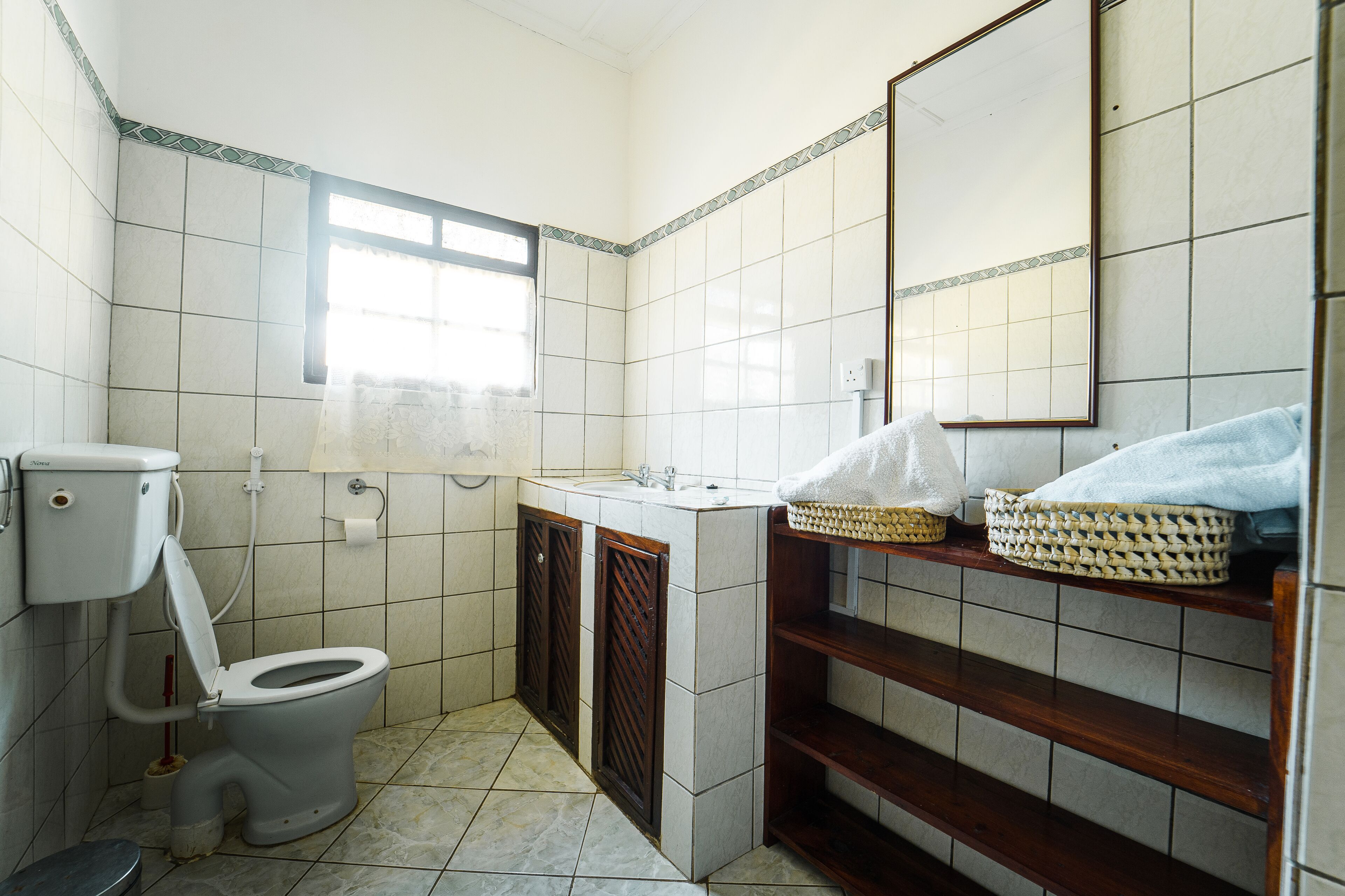 Ein kleines Badezimmer mit Toilette, Waschbecken und Spiegel. Ein Fenster sorgt für natürliches Licht, und ein Regal mit Körben ist ebenfalls vorhanden.