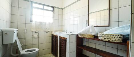 A small bathroom with a toilet, sink, and mirror. There is a window providing natural light and a shelf with baskets.