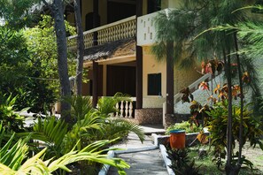 A pathway leading to a building with a thatched roof balcony, surrounded by lush greenery and a stone wall. - GK Palm Resort Watamu (Watamu)