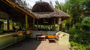 A covered outdoor seating area with a thatched roof, bar stools, and a wooden table. - GK Palm Resort Watamu (Watamu)