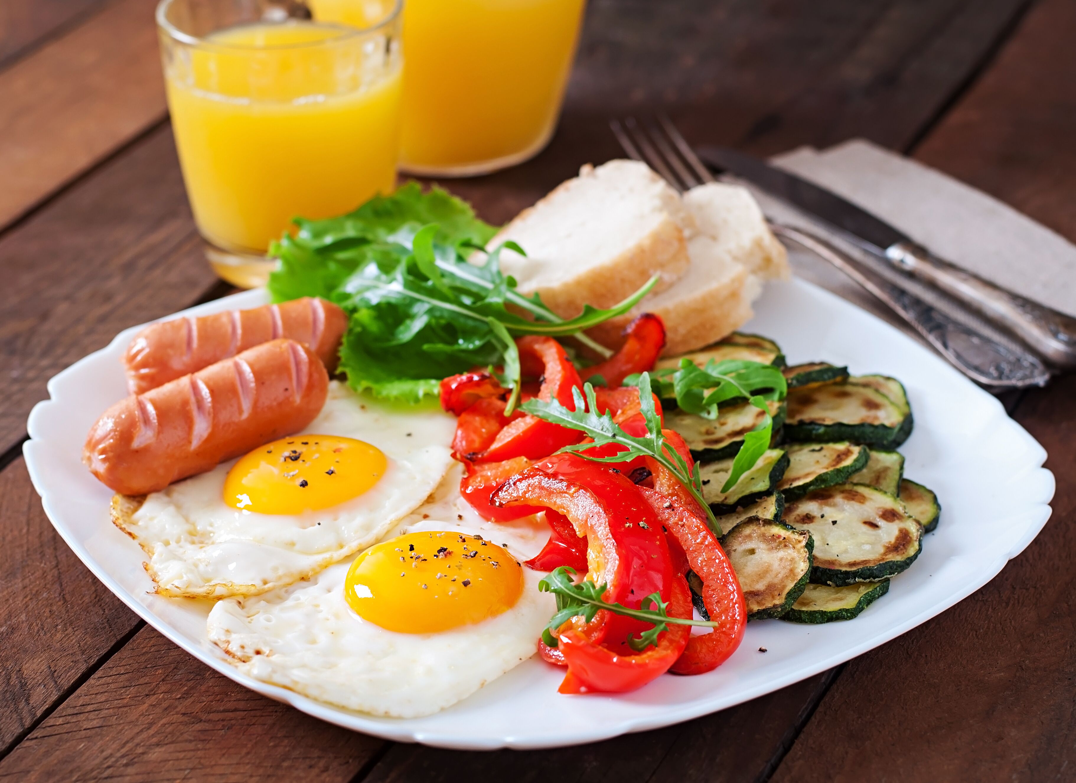 A plate of food with eggs, sausages, peppers, and zucchini slices, accompanied by a glass of orange juice and a napkin with utensils.