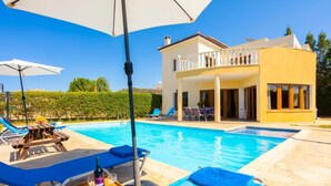 Zona de piscina con sillones reclinables, una mesa con una botella y vasos, y un edificio con balcón.