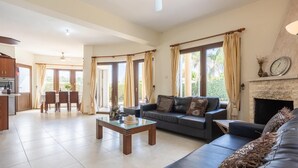 A spacious living area with a sofa, a coffee table, and a fireplace. There is a dining area with a table and chairs, and large windows with curtains. - Villa Tsikkos Ena - Three Bedroom Villa, Sleeps 6 (Argaka)