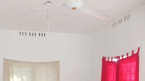 Un salon avec deux canapés sombres, une table basse en bois, un ventilateur de plafond et une fenêtre ornée de rideaux rouges.