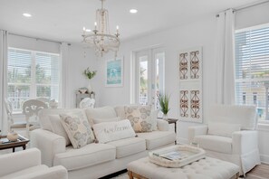 Uma sala de estar espaçosa com um sofá branco, uma poltrona combinando e uma mesa de centro. Um lustre pendurado no teto e várias obras de arte emolduradas nas paredes.