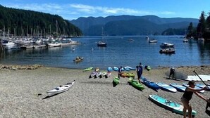 A beach with kayaks and paddleboards, boats in the water, and mountains in the background. - Rustic modern garden level suite (North Vancouver)