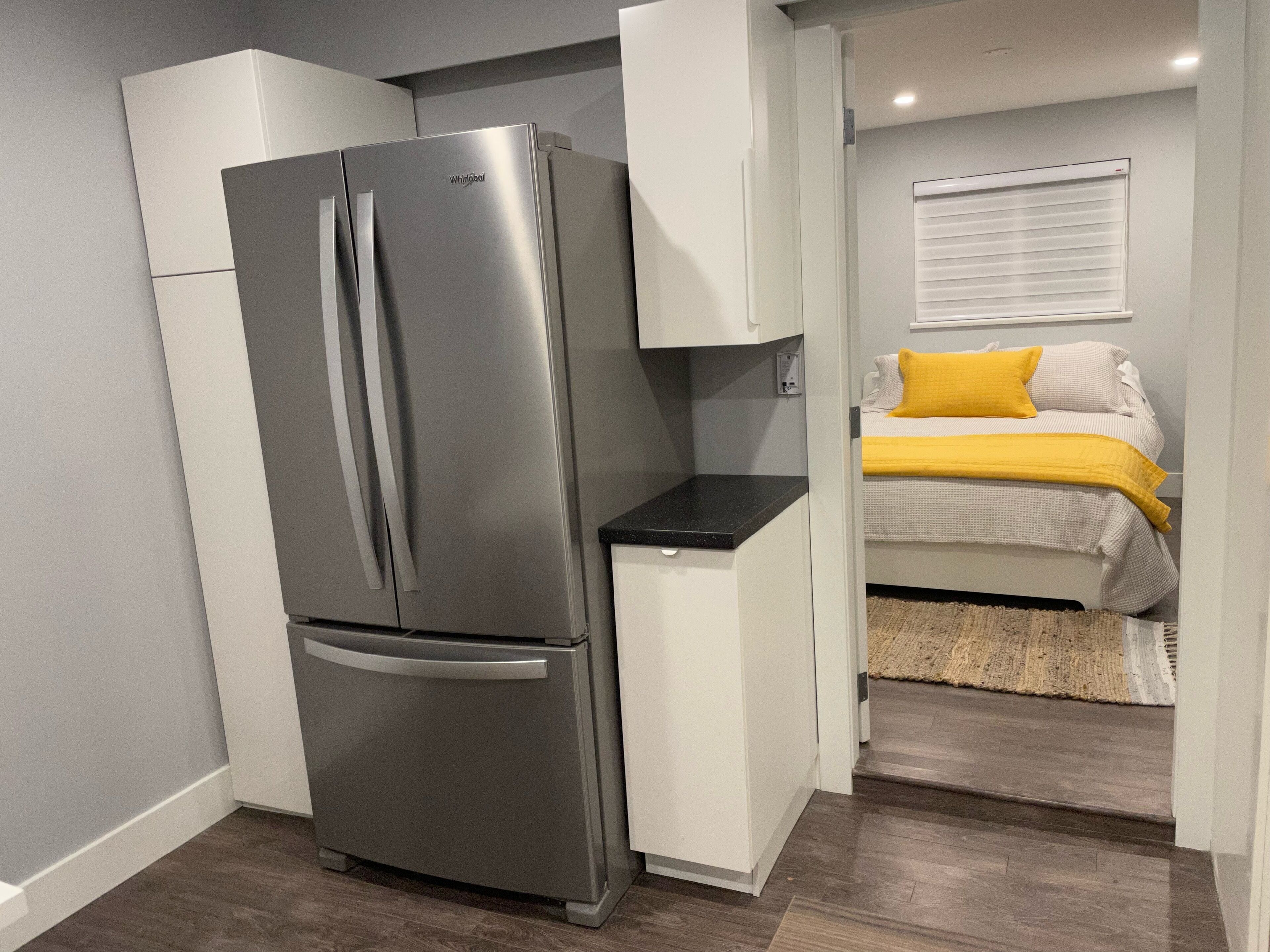 A compact kitchen with a stainless steel refrigerator, white cabinetry, and a bed with a yellow pillow in the adjacent room.