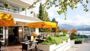 An outdoor seating area with yellow umbrellas, a stone planter with greenery, and a view of a lake and mountains. - Rustic modern garden level suite (North Vancouver)