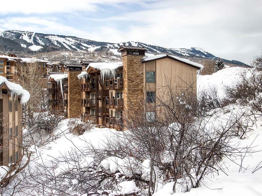 A snow-covered building with stone and wood exterior, surrounded by snow-covered trees and mountains in the background.