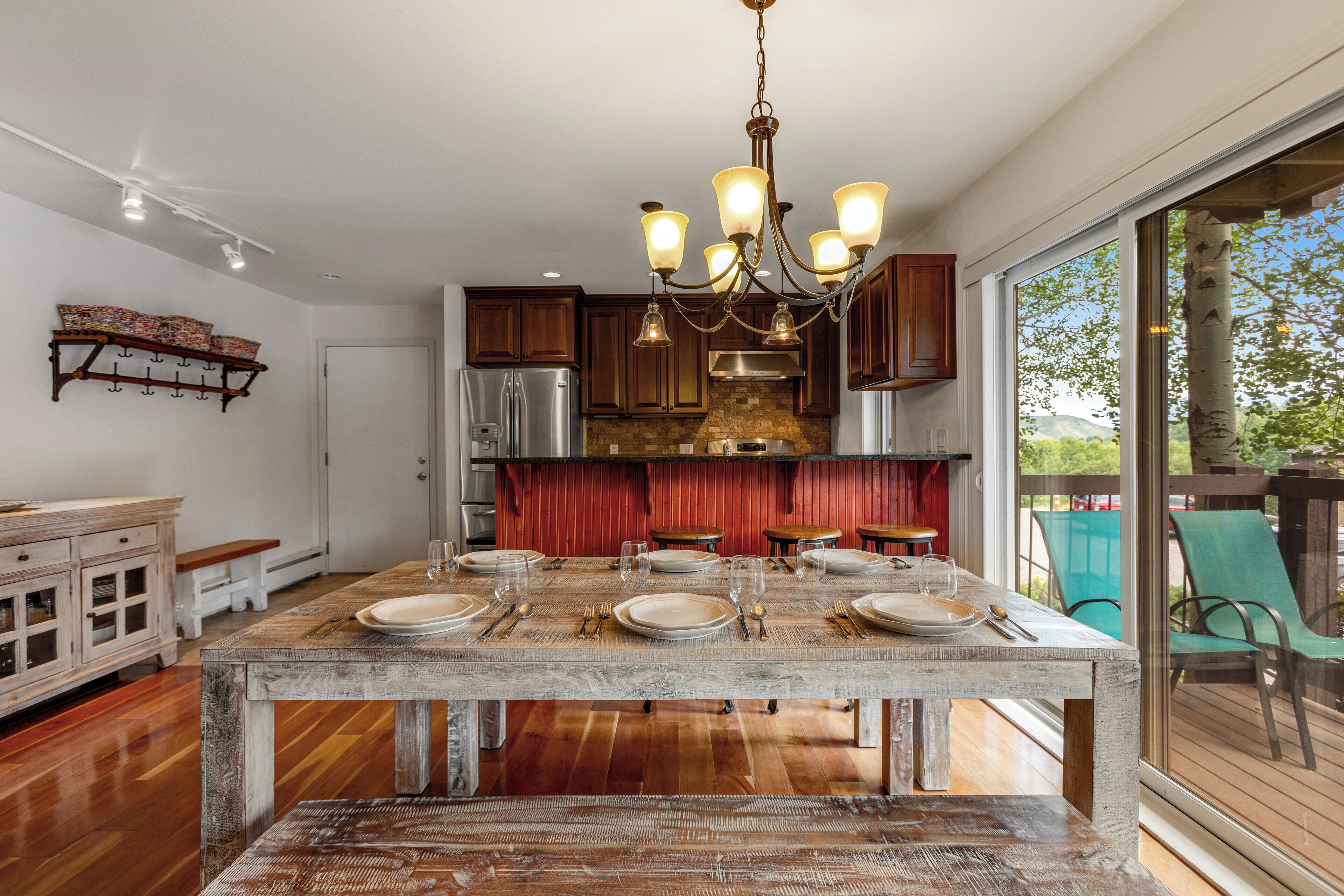 Un coin repas avec une table en bois rustique dressée pour six personnes, un lustre et une cuisine dotée de meubles en bois et d’un mur d’accent rouge.