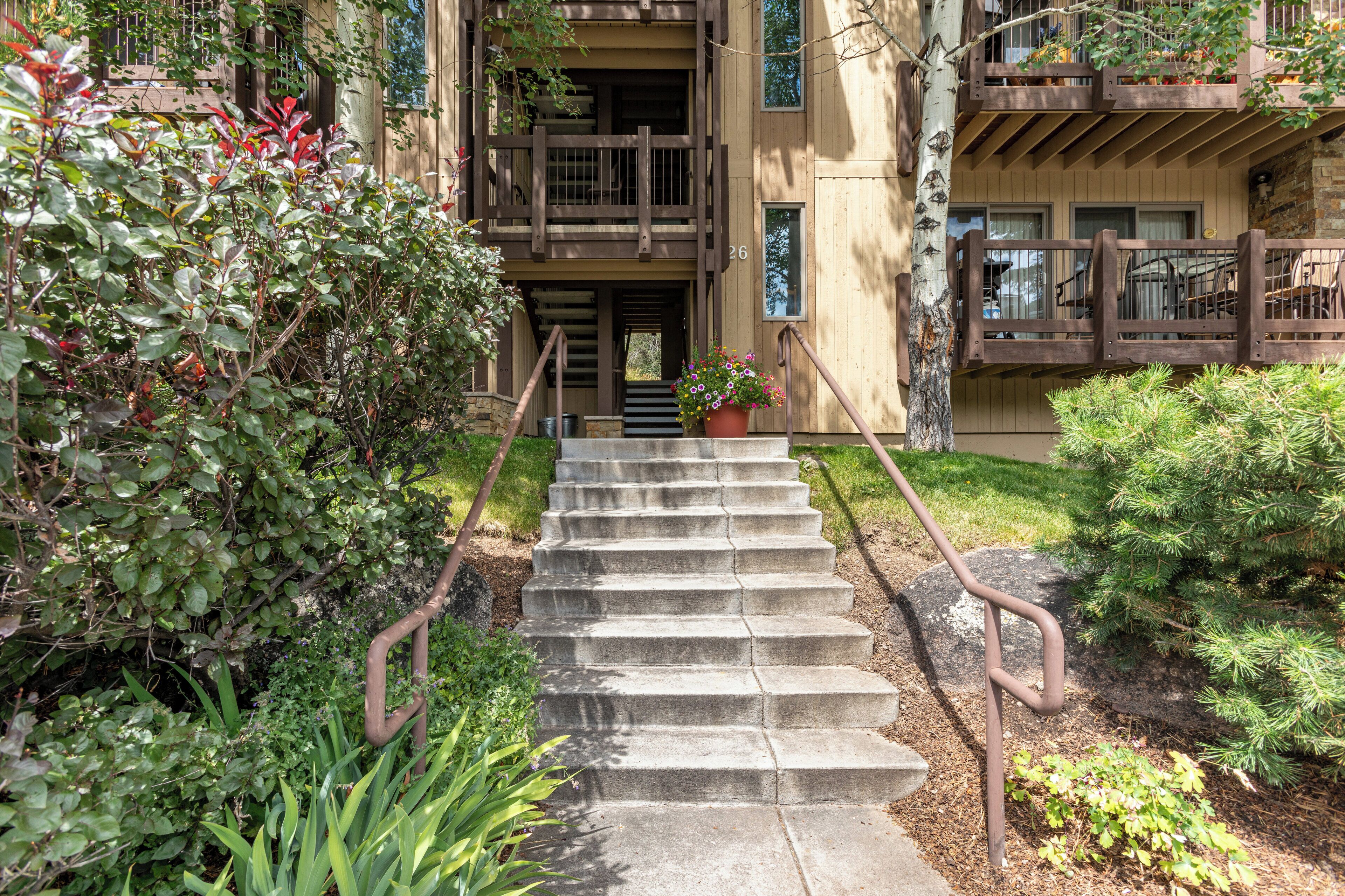 A wooden staircase leading to a building with multiple balconies, surrounded by lush greenery and a potted plant at the bottom.