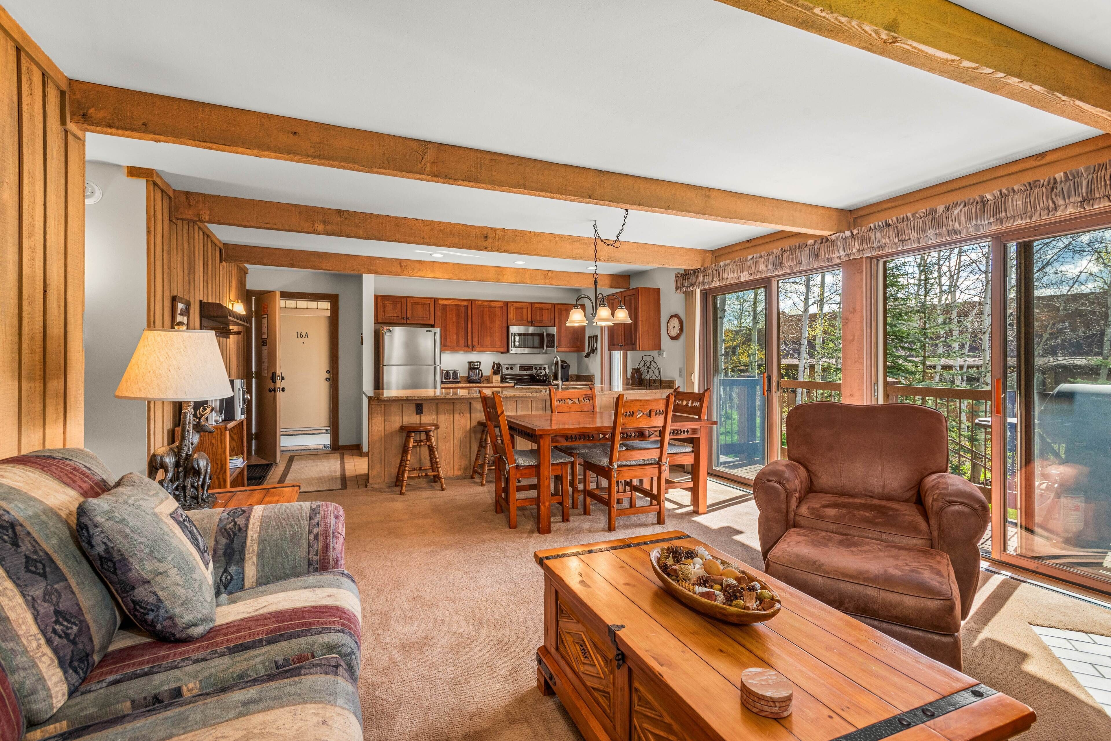 A cozy living room with a sofa, a wooden coffee table, and a brown leather chair. A kitchen with wooden cabinets and a dining area with a table and chairs is visible in the background.