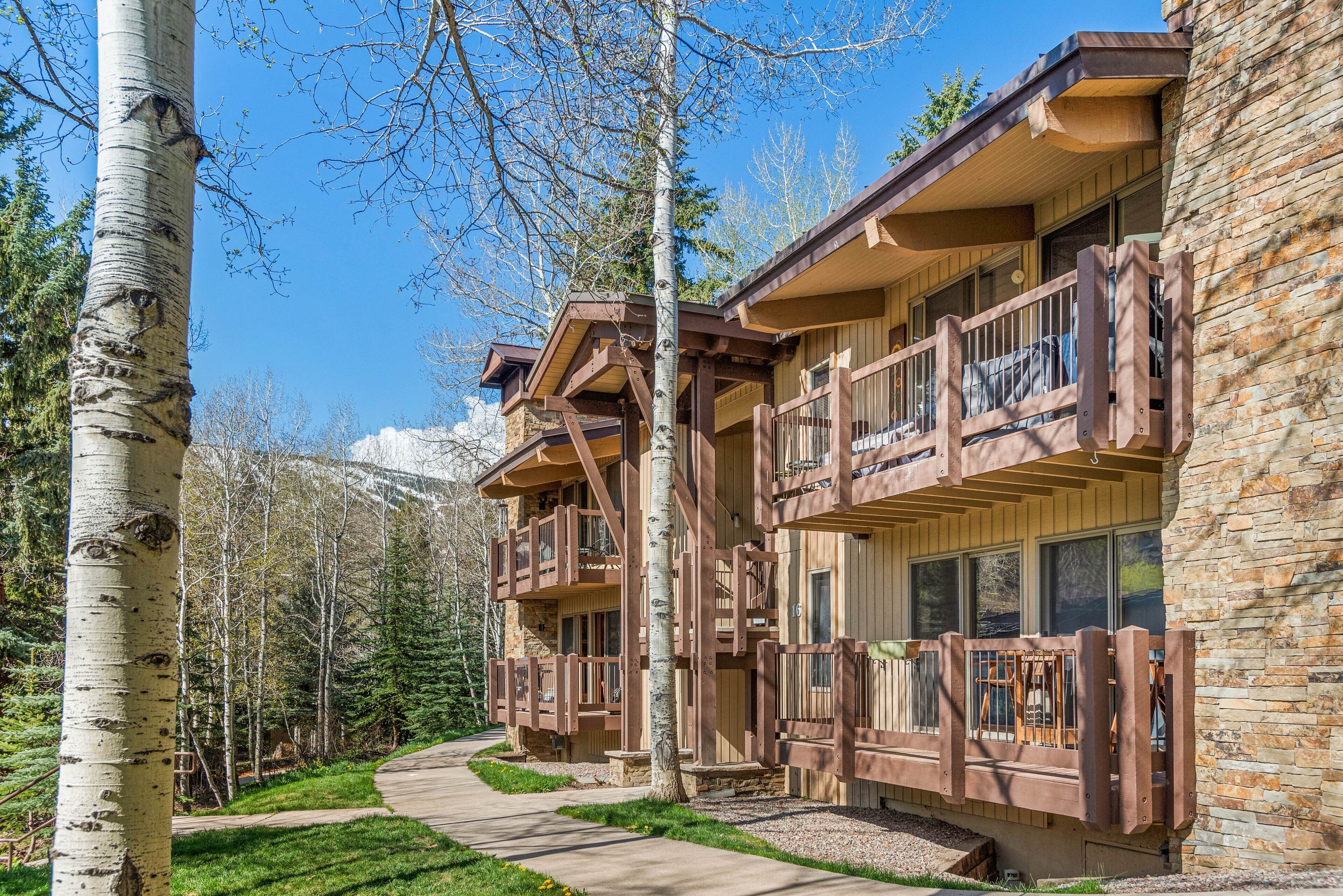 A multi-story lodge with wooden balconies and stone walls, surrounded by trees and a clear sky.