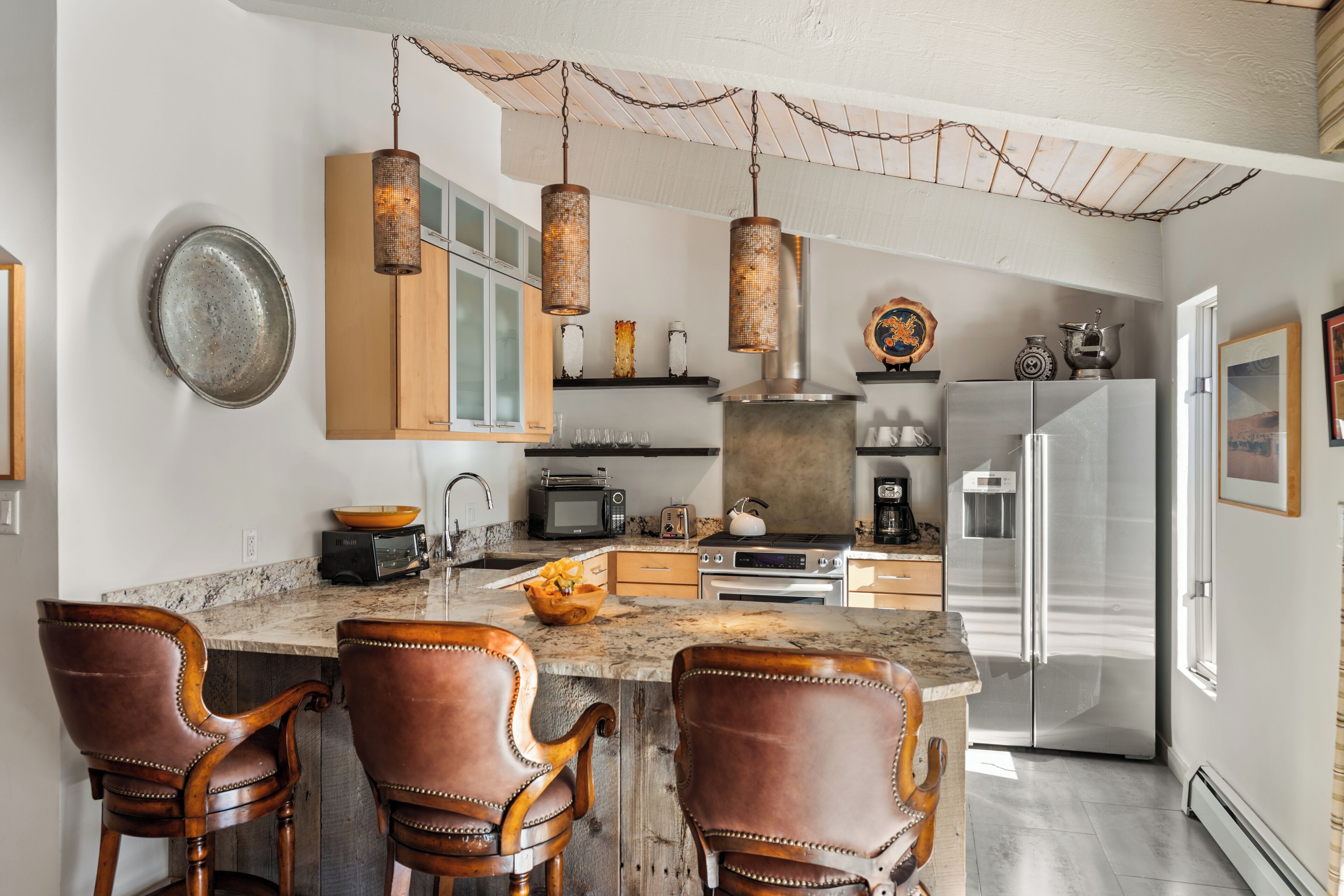 A kitchen with a marble countertop, wooden chairs, a stainless steel refrigerator, and hanging pendant lights.
