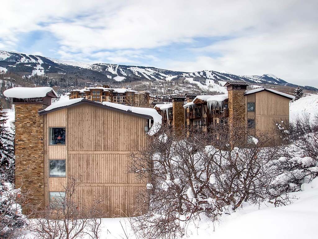 Ein schneebedecktes Gebäude mit Holzfassade und zahlreichen Fenstern, umgeben von schneebedeckten Bäumen und Bergen im Hintergrund.