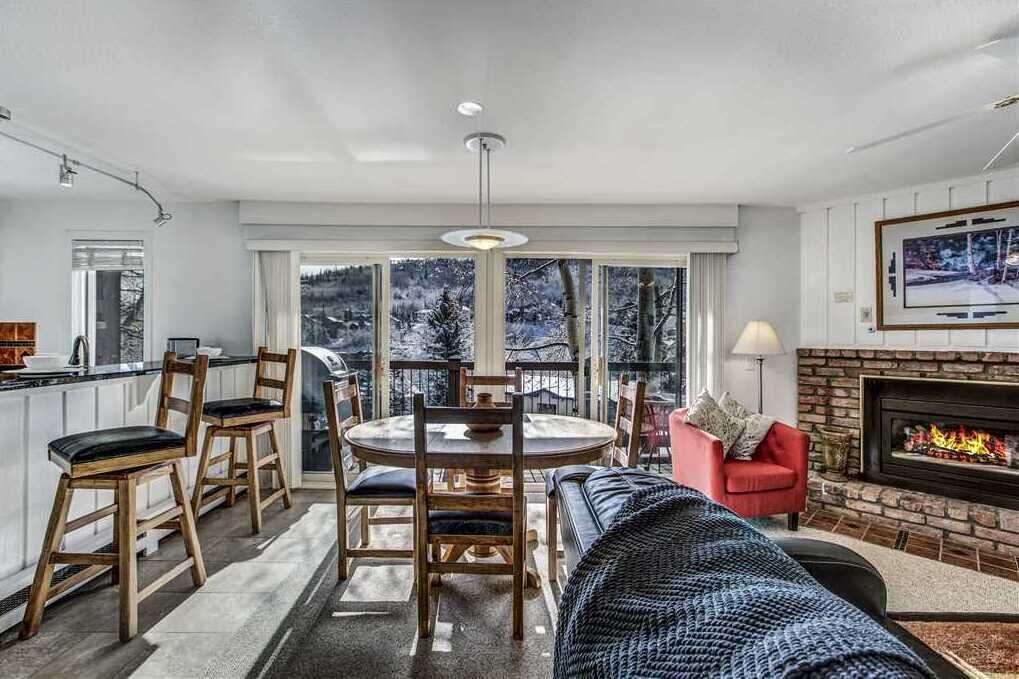 A modern living room with a fireplace, a dining area with wooden chairs, and a large window offering a view of trees.