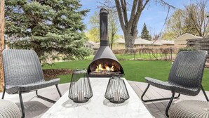 An outdoor seating area with two wicker chairs and a table, a metal fire pit with flames, and a backdrop of trees and houses. - Benelo | Midcentury Modern Single-Level Home, 3 BR (Sioux Falls)