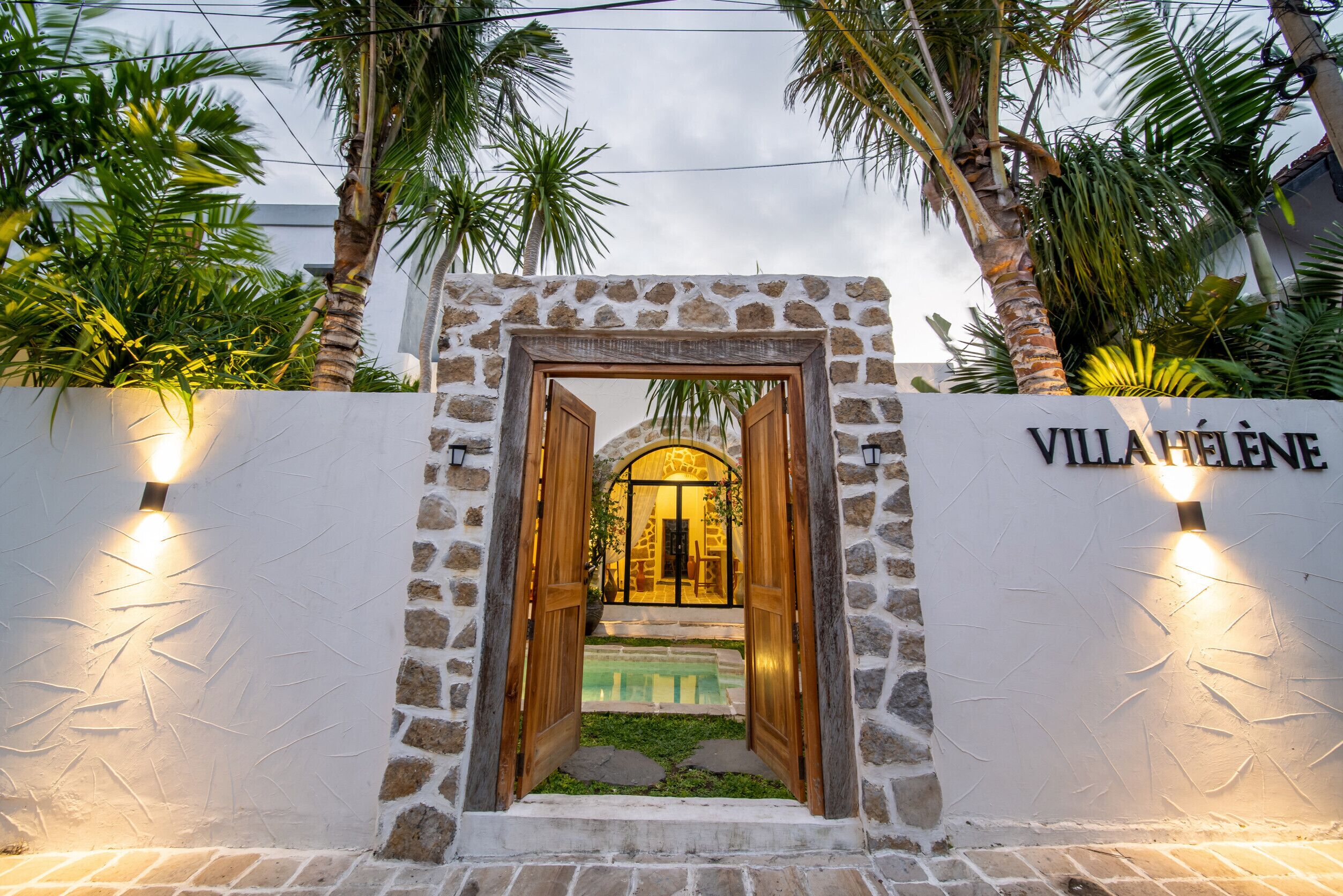 Entrada de una villa con un muro de piedra, puertas de madera y una zona de piscina visible a través del arco.