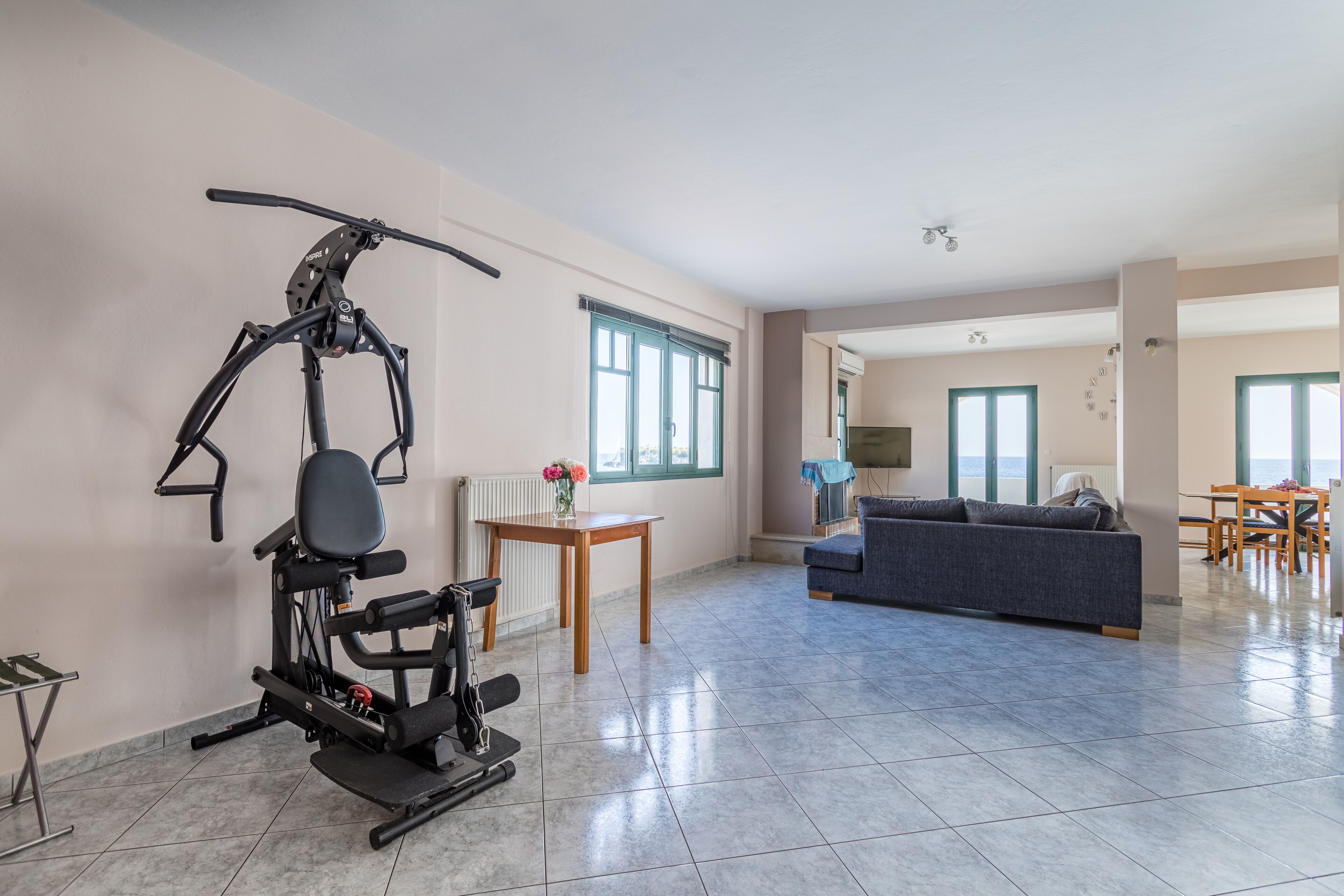 A spacious living area with a stationary exercise bike, a sofa, a dining table, and a view of the outside through large windows.
