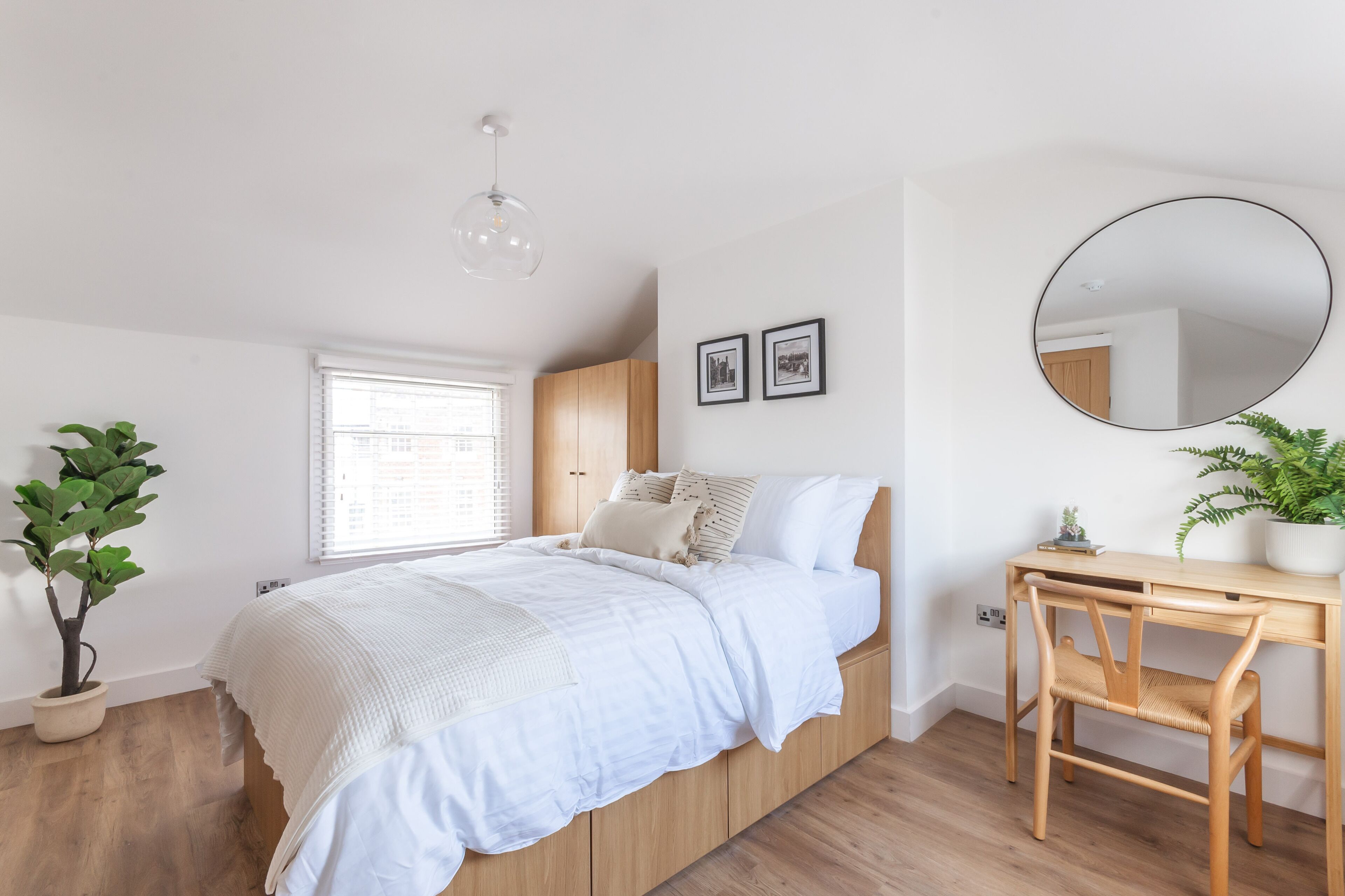 A bedroom with a bed, a desk, a chair, a potted plant, a window, and a mirror.