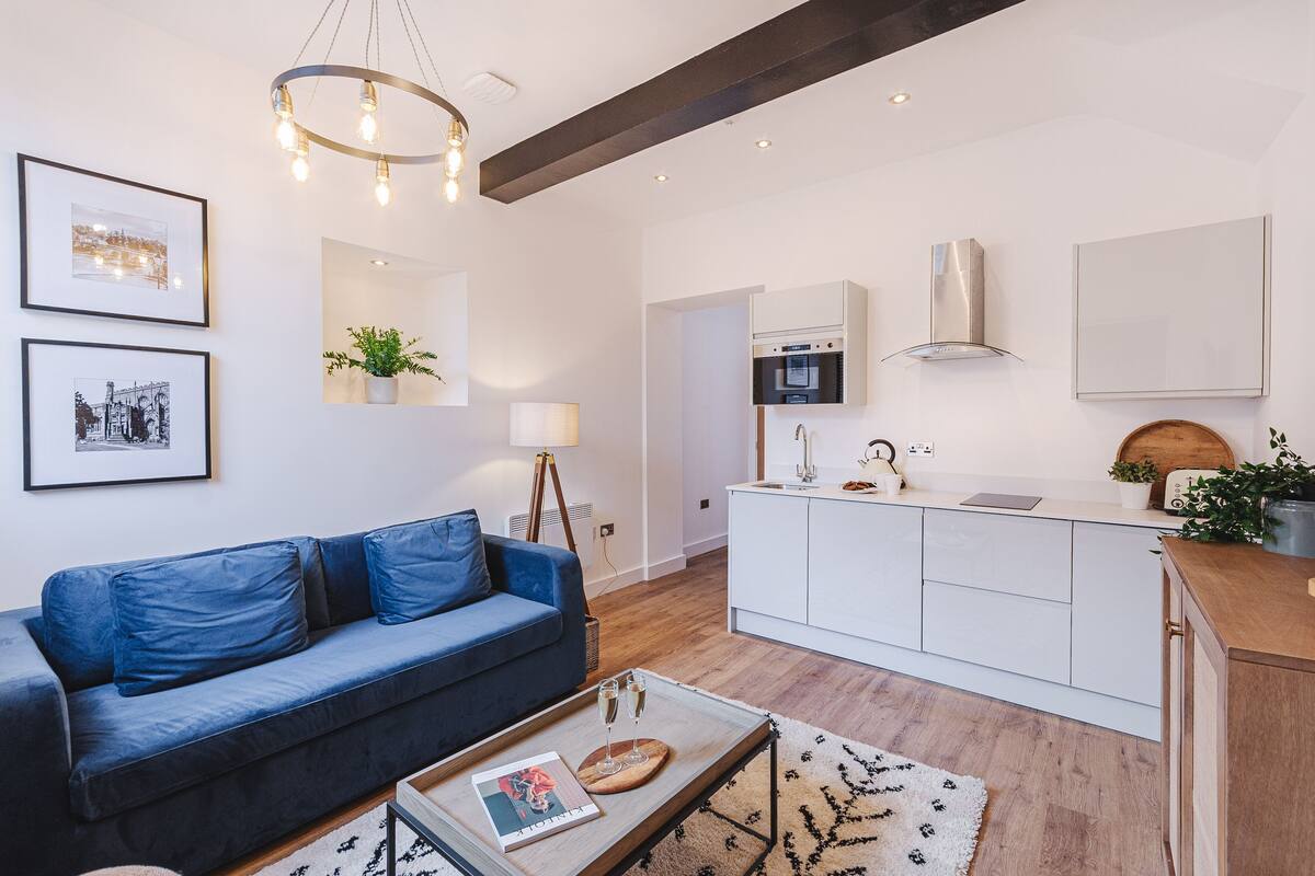 Een moderne woonkamer met een blauwe bank, een salontafel met tijdschriften, een kroonluchter, en een keuken met witte kastjes en een fornuis.