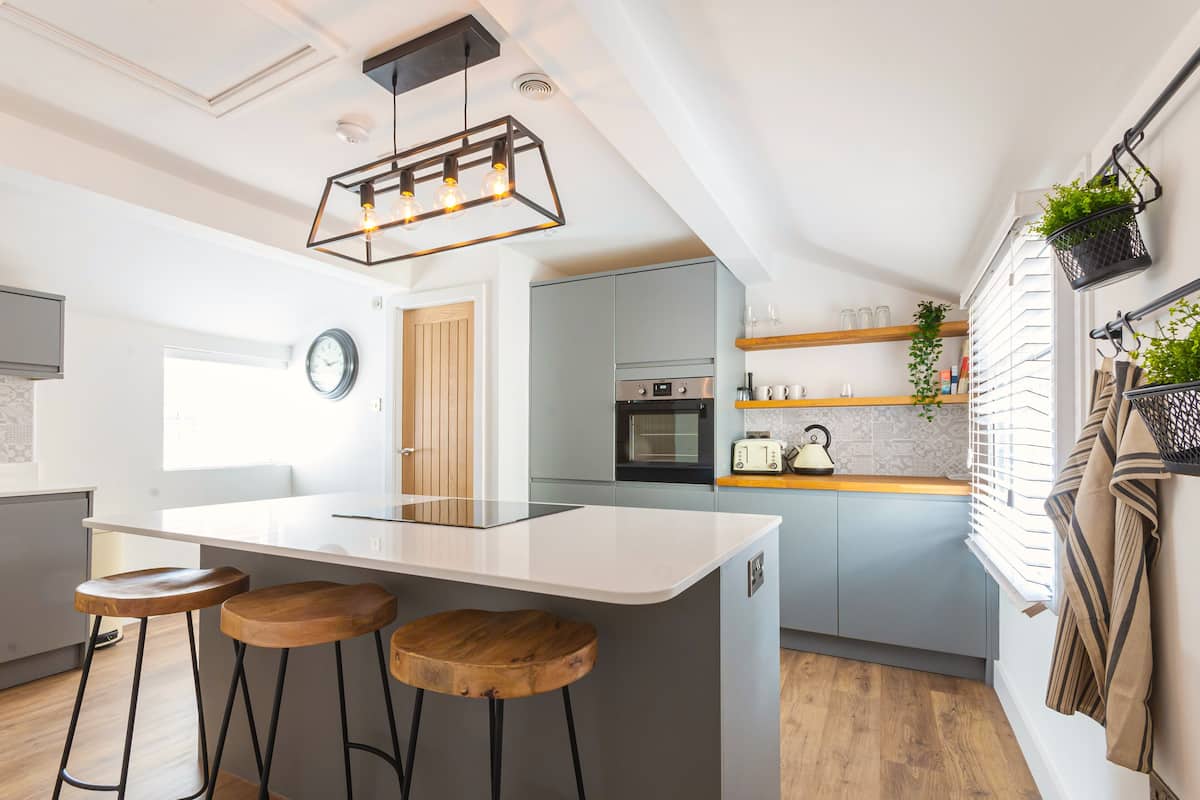 Een moderne keuken met een centraal kookeiland, houten krukken, een hanglamp en een raam met zonwering.