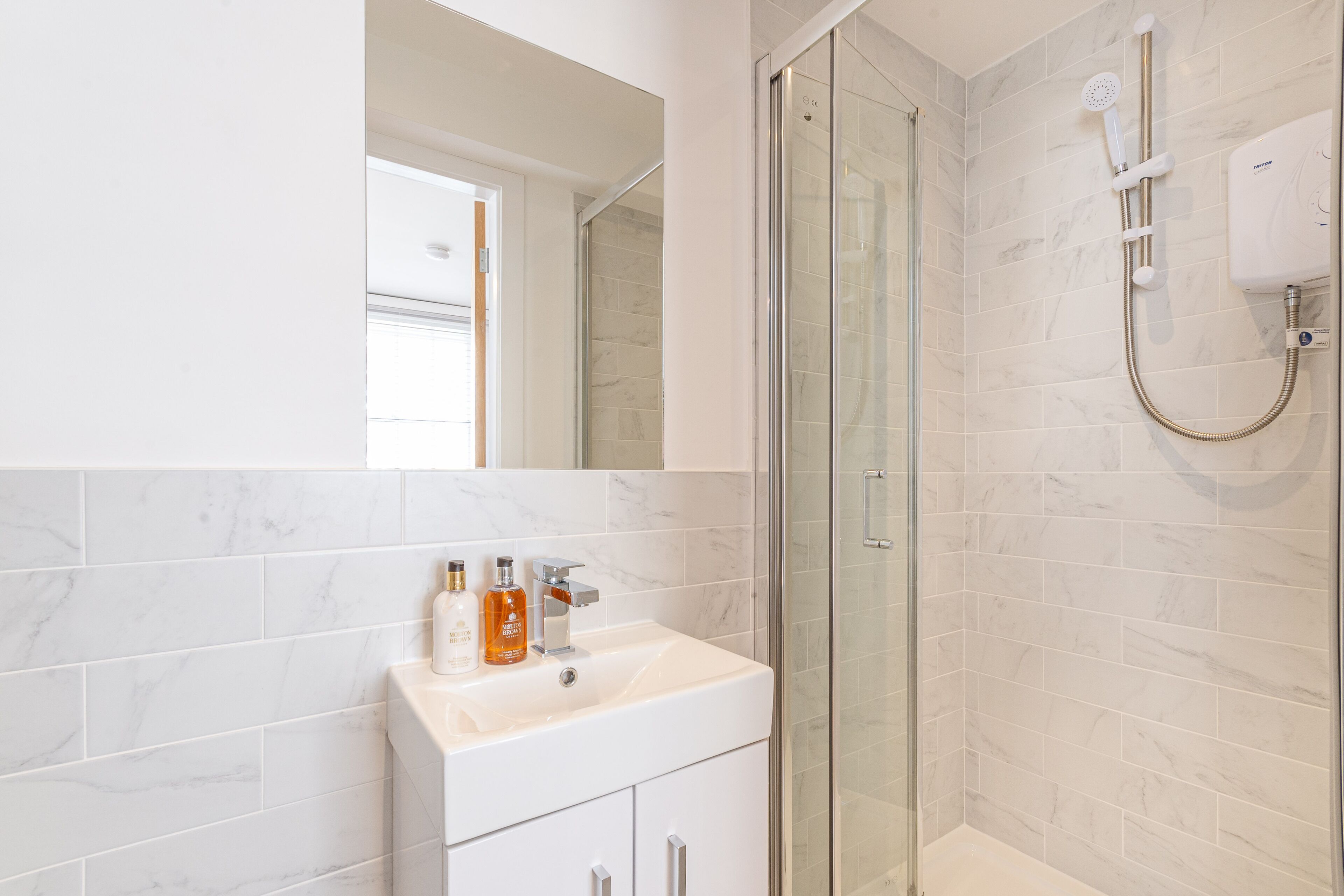 Uma casa de banho moderna com chuveiro em cabine de vidro, lavatório branco com produtos de higiene e azulejos de mármore.
