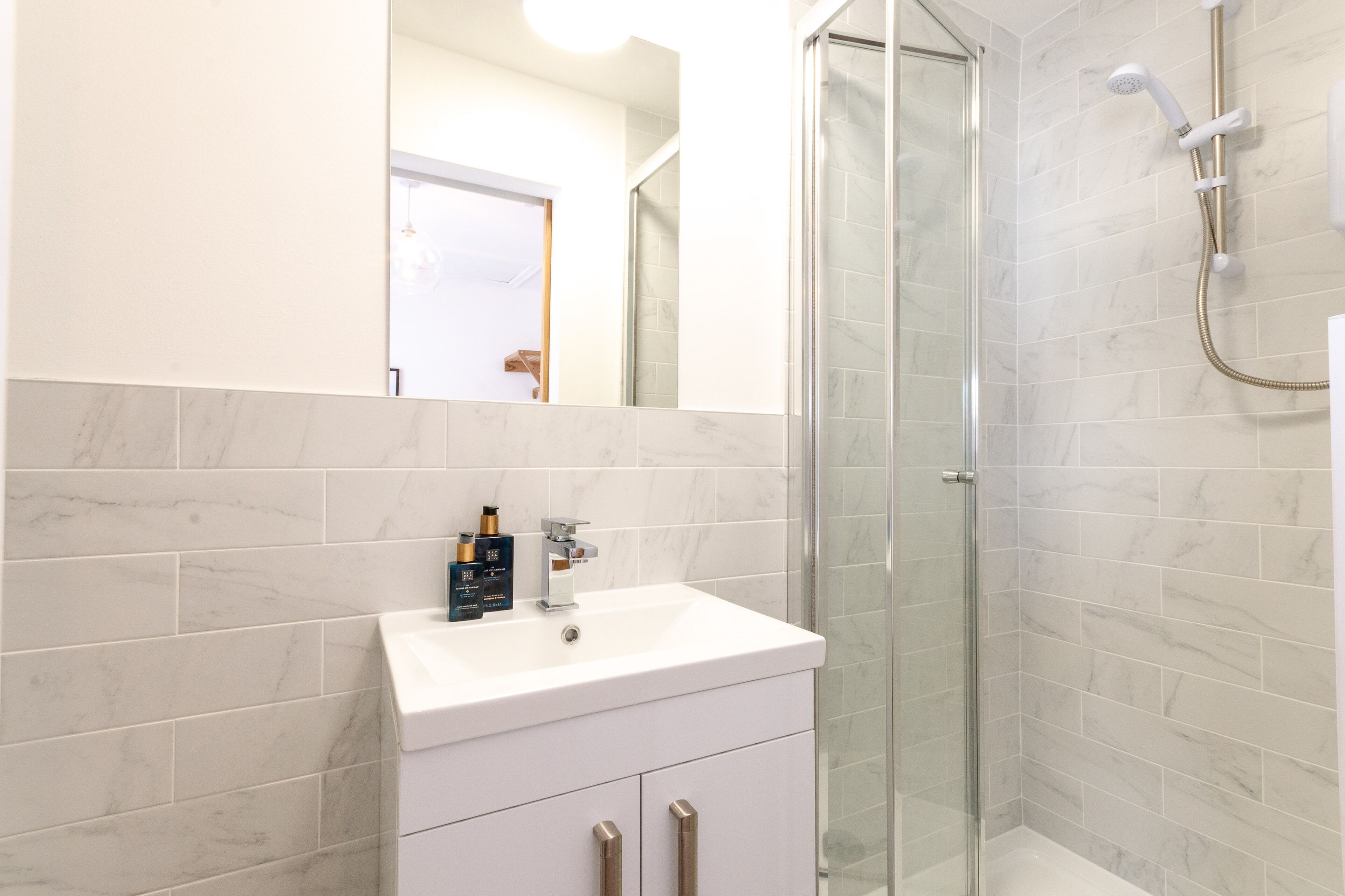 A modern bathroom with a white sink, marble tiles, a glass shower enclosure, and a mirror above the sink.