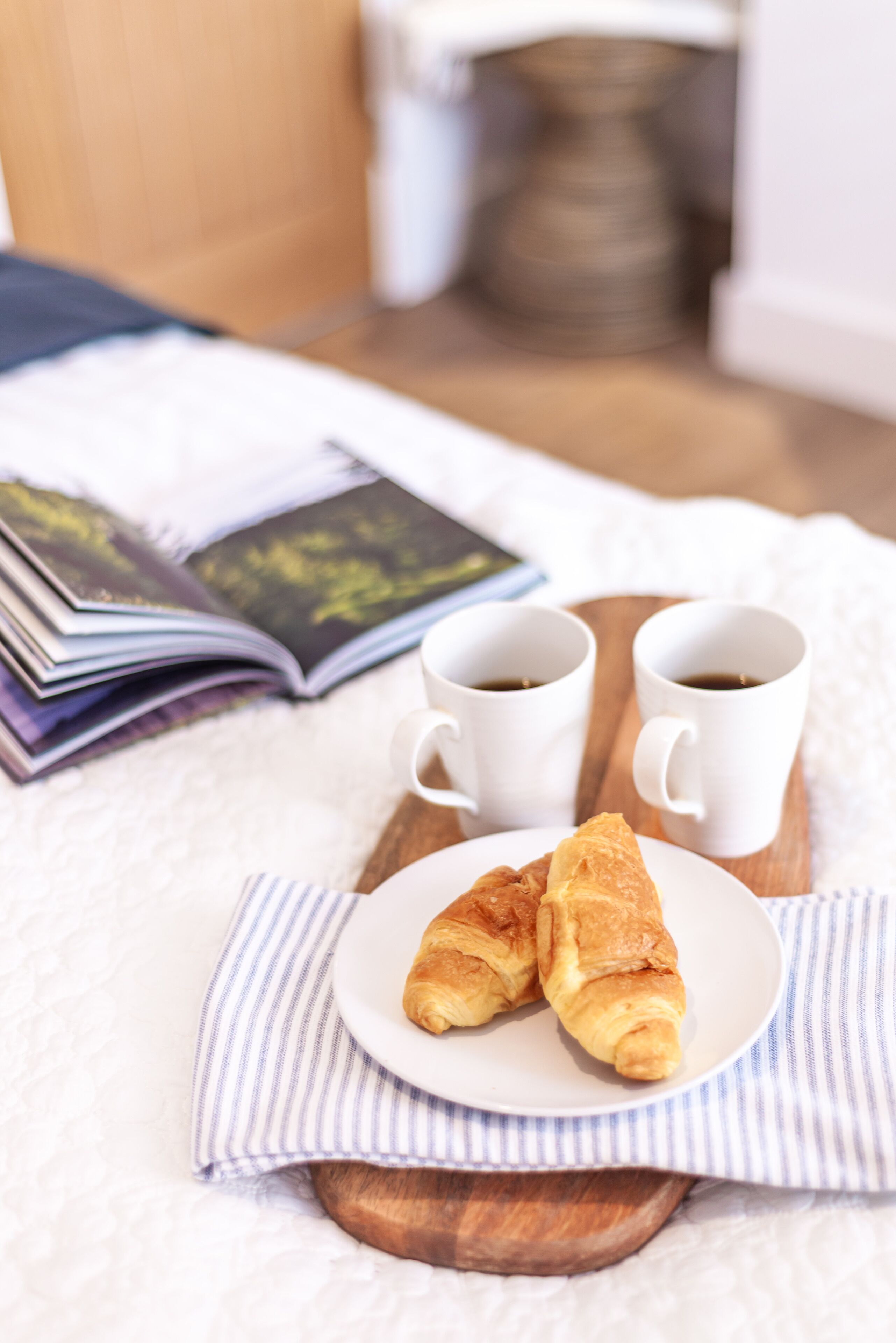 Un desayuno con cruasanes en un plato, dos tazas de café y una revista abierta sobre una bandeja de madera.