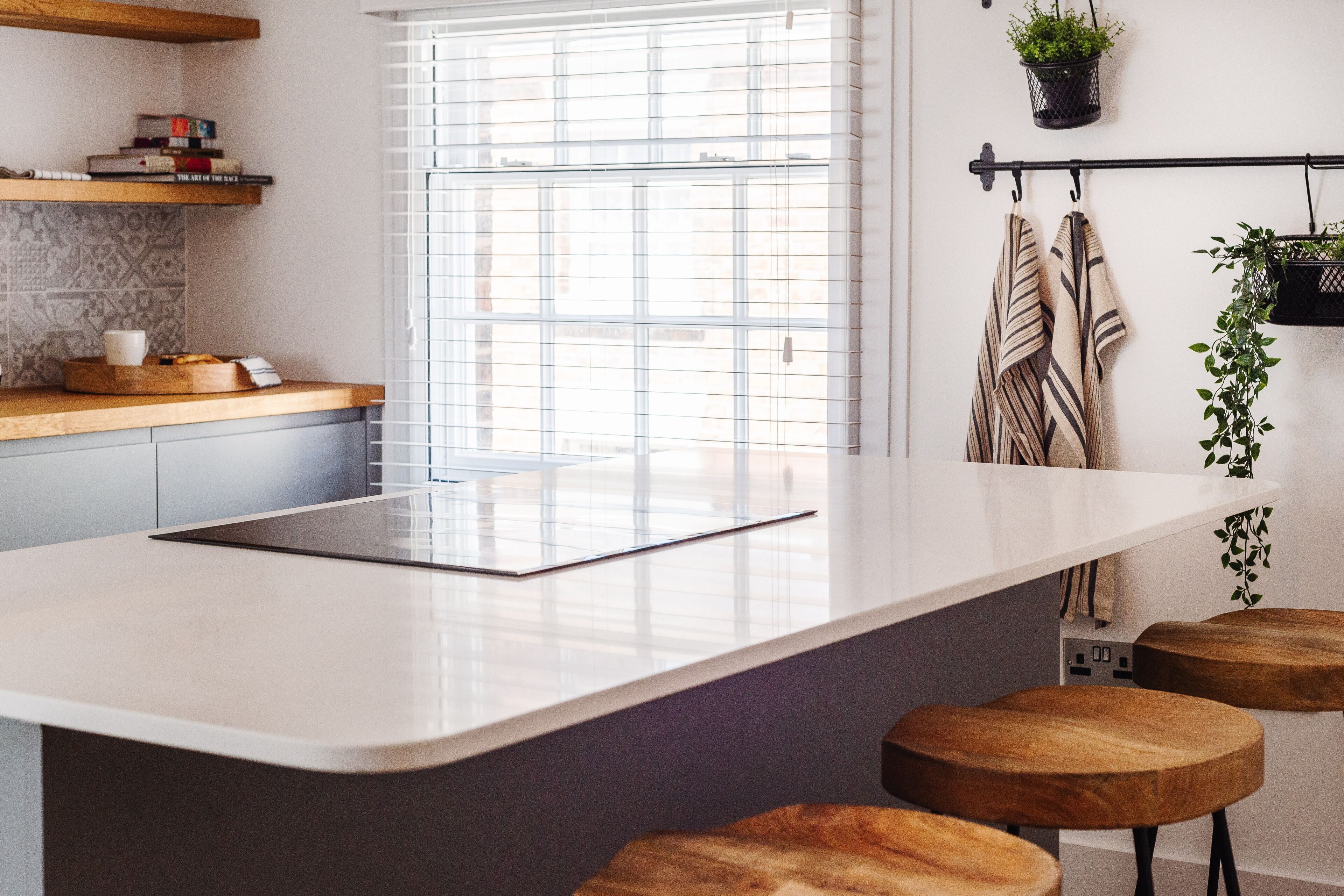 A modern kitchen with a central island, wooden stools, a window with blinds, and a hanging plant.