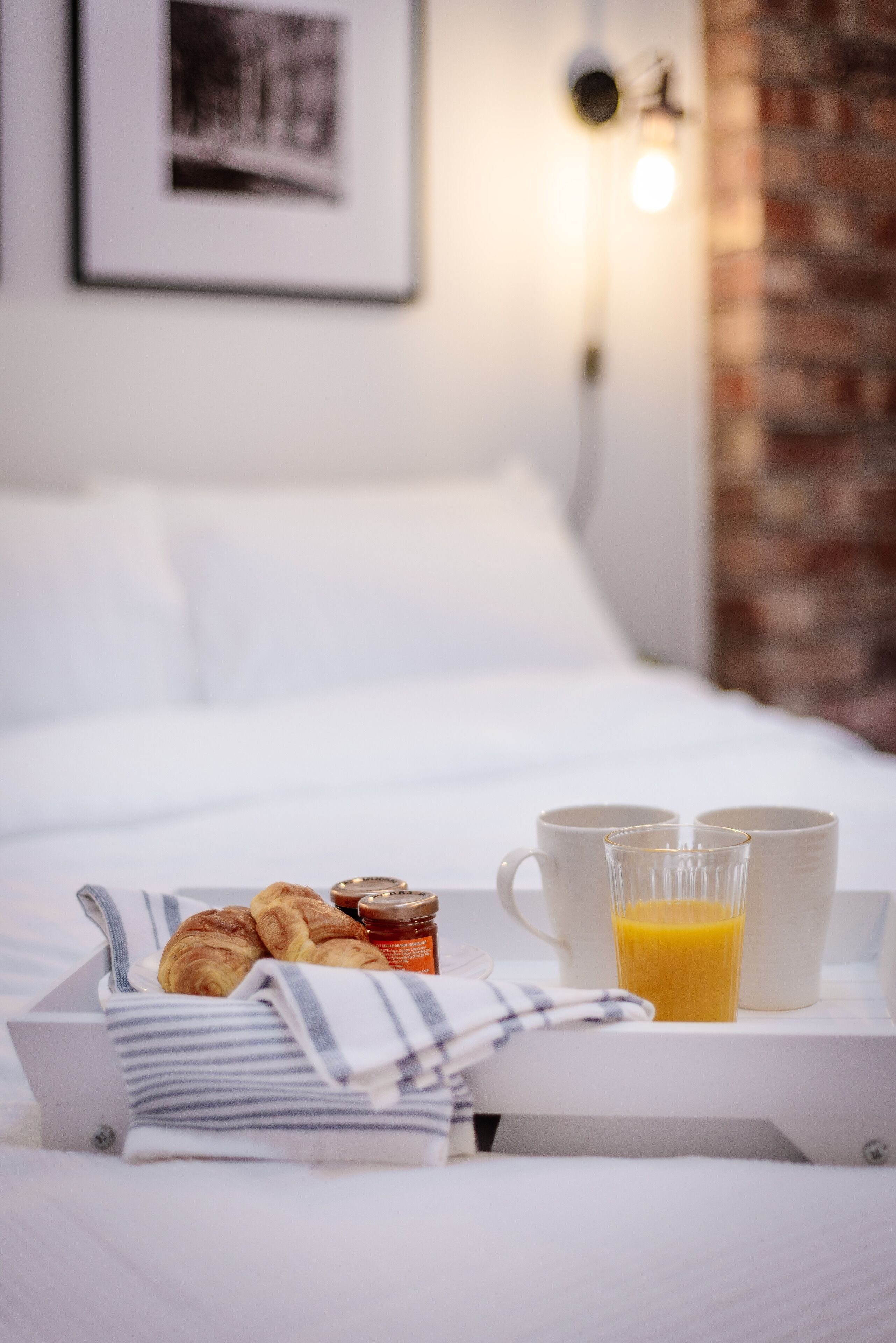 A neatly arranged breakfast tray with croissants, jam, and orange juice on a bed with white linens and a brick wall in the background.